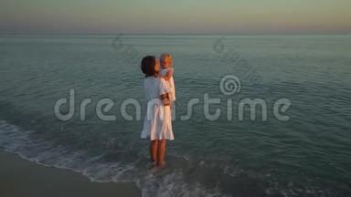日落时分，年轻漂亮的女人和她的小女儿在海滨散步。 妈妈把女儿抱在怀里。 他们